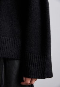 Musta neulottu neule löysällä istuvuudella, jossa on resoroidut hihansuut ja pehmeä tekstuuri, kuvassa käsi hihalla.