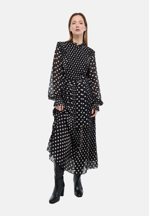 Femme portant une robe midi noire à pois blancs, manches longues transparentes, volants et ceinture à la taille, associée à des bottes noires à talons.