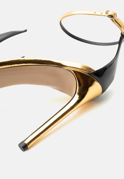 Μαύρο και χρυσό σανδάλι με ψηλό τακούνι, λουράκι αστραγάλου, μυτερή μύτη τακουνιού και γυαλιστερό φινίρισμα σε λευκό φόντο.