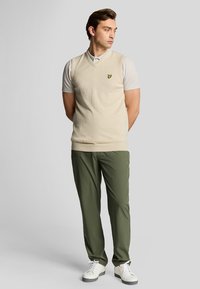 Maglione beige con collo a V sopra una polo grigia chiara, abbinato a pantaloni verde oliva e sneakers bianche. Piccolo logo giallo sul maglione.