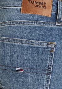 Jean en denim bleu avec une patch en cuir portant l'étiquette "TOMMY JEANS", dotée de coutures contrastantes et d'un petit logo brodé en rouge, blanc et bleu.