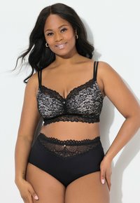 Lachende vrouw met lang donker haar, die een zwarte kanten bralette en bijpassende hoge kanten slipjes draagt tegen een witte achtergrond.