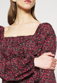 Musta kukkamekko puffihihoilla, jossa on neliönmuotoinen pääntie ja pieniä vaaleanpunaisia ja valkoisia kukkakuoseja. Teksturoitu kangas, jossa on rypytetty yksityiskohta.