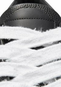Black leather sneaker with thick white laces covering the upper. Close-up of the textured surface and stitching details.