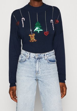 Pull bleu marine avec des ornements de Noël suspendus en sequins : cœurs, cannes en sucre d'orge, rennes et sapin, porté avec un jean bleu clair.
