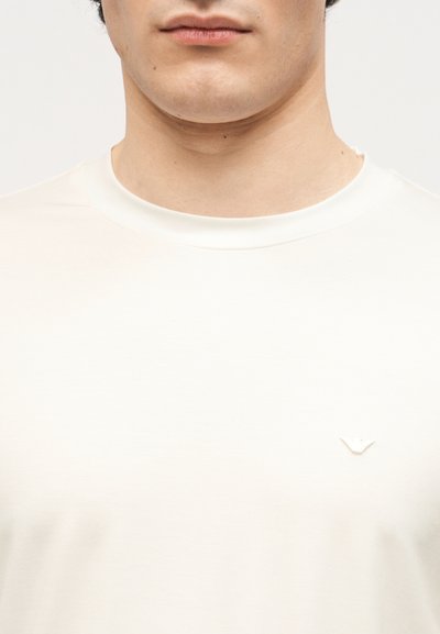 Κοντινή λήψη ενός ατόμου που φοράει ένα απλό λευκό μπλουζάκι με στρογγυλή λαιμόκοψη και μικρό λευκό λογότυπο στο αριστερό στήθος.