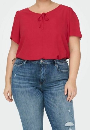 Person trägt eine rote Kurzarmbluse mit vorderer Schleifendetail und blaue, hoch taillierte Jeans mit Used-Look vor einfarbigem Hintergrund.