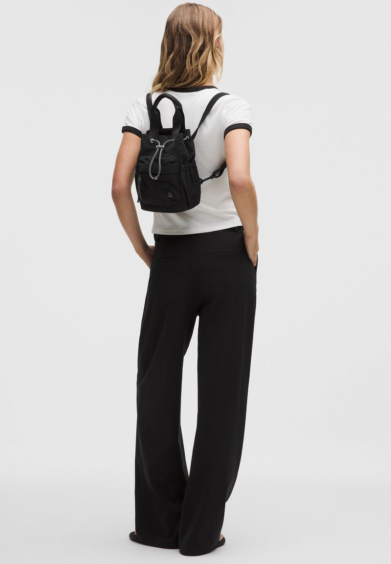 Frau trägt weißes Hemd und schwarze Hose mit kleinem schwarzem Rucksack mit Kordelzug, steht mit den Händen in den Taschen, Blick nach hinten gewandt.