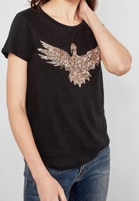 Czarna koszulka z krótkim rękawem z aplikacją w postaci cekinowego orła w kolorze różowego złota z przodu. Materiał wydaje się być miękki i ma luźny krój.