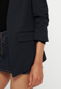 Femme portant un blazer foncé avec manches retroussées sur un top en dentelle noir et un short en denim foncé, bras détendu le long du corps.