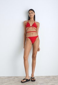 Red bikini with triangle top and crisscross straps. The bottom features a low-rise cut. Model wearing black sandals, standing on a stone floor.
