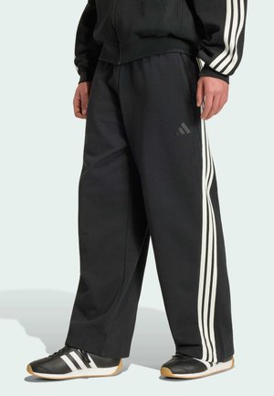 Personne portant un pantalon de survêtement Adidas noir avec des bandes blanches sur les côtés et des baskets noires assorties avec des bandes blanches.