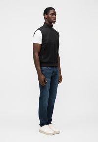 Μαύρο αμάνικο γιλέκο με ψηλό γιακά, φερμουάρ στο μπροστινό μέρος και πλαϊνές τσέπες, συνδυασμένο με λευκό μπλουζάκι και μπλε τζιν, φορεμένο με λευκά αθλητικά παπούτσια.