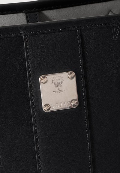 Sac en cuir noir avec une plaque métallique argentée gravée du logo MCM München et du numéro de série V-8743.