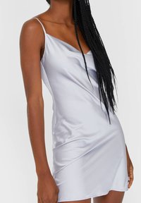 Robe nuisette en satin argenté avec de fines bretelles et un design drapé délicat. Le tissu a une texture lisse et brillante.