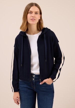 Young woman wearing a navy zip-up hoodie with white stripes on sleeves, white t-shirt, and dark blue jeans, standing against a beige background.