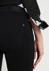 Jeans de mezclilla negra con un bolsillo trasero que presenta costuras curvas y un pequeño botón de metal. El tejido parece suave y sin signos visibles de uso.
