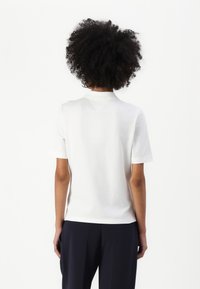 Personne aux cheveux bouclés portant un t-shirt blanc uni à manches courtes et un pantalon sombre, debout dos à un fond blanc.
