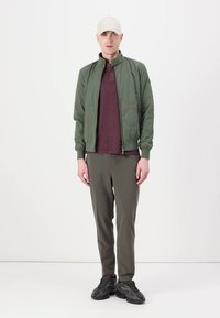 Zielona pikowana kurtka bomber, burgundowy polo z fakturą, oliwkowe spodnie i ciemne trampki. Model stoi przodem do kamery na neutralnym tle.