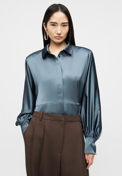 Femme aux longs cheveux foncés portant un chemisier en satin bleu à manches bouffantes et un pantalon marron taille haute, les mains partiellement dans les poches, fond neutre.