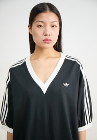 Melna oversized T-krekls ar baltu V veida izgriezumu un baltām svītrām uz piedurknēm. Neliels Adidas logo uz krūtīm. Mīksta auduma tekstūra.