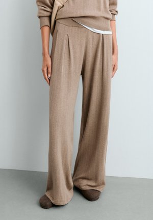 Personne portant un pantalon beige à rayures fines, ample à taille haute avec des plis devant et des chaussures plates marron, debout contre un mur gris.