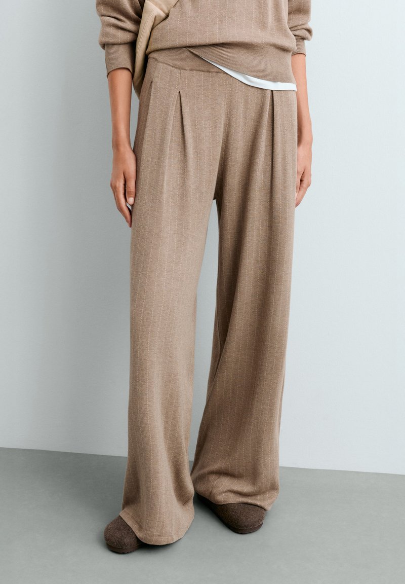 Personne portant un pantalon beige à rayures fines, ample à taille haute avec des plis devant et des chaussures plates marron, debout contre un mur gris.