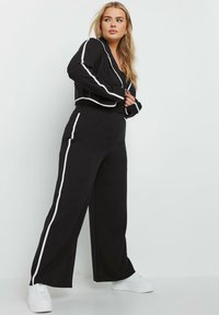Czarny zestaw odzieży domowej składający się z krótkiej kurtki z białymi akcentami oraz spodni z szerokimi nogawkami i bocznymi paskami. Materiał wydaje się gładki i elastyczny.