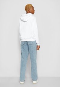 Felpa bianca con cappuccio e cordino, abbinata a jeans oversize azzurri chiari. I jeans presentano una texture sbiadita e tasche posteriori, indossati con scarpe bianche.