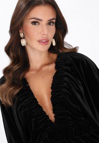 Fekete bársonyruha összegyűrt nyakkivágással és hosszú ujjakkal. Arany textúrájú fülbevalókkal és finom nyaklánccal viselve.