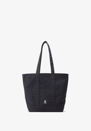 Zwarte canvas tote bag met twee lange handvatten, een voorvak en een klein logo-label nabij de onderkant. Eenvoudig ontwerp, ruime vorm.