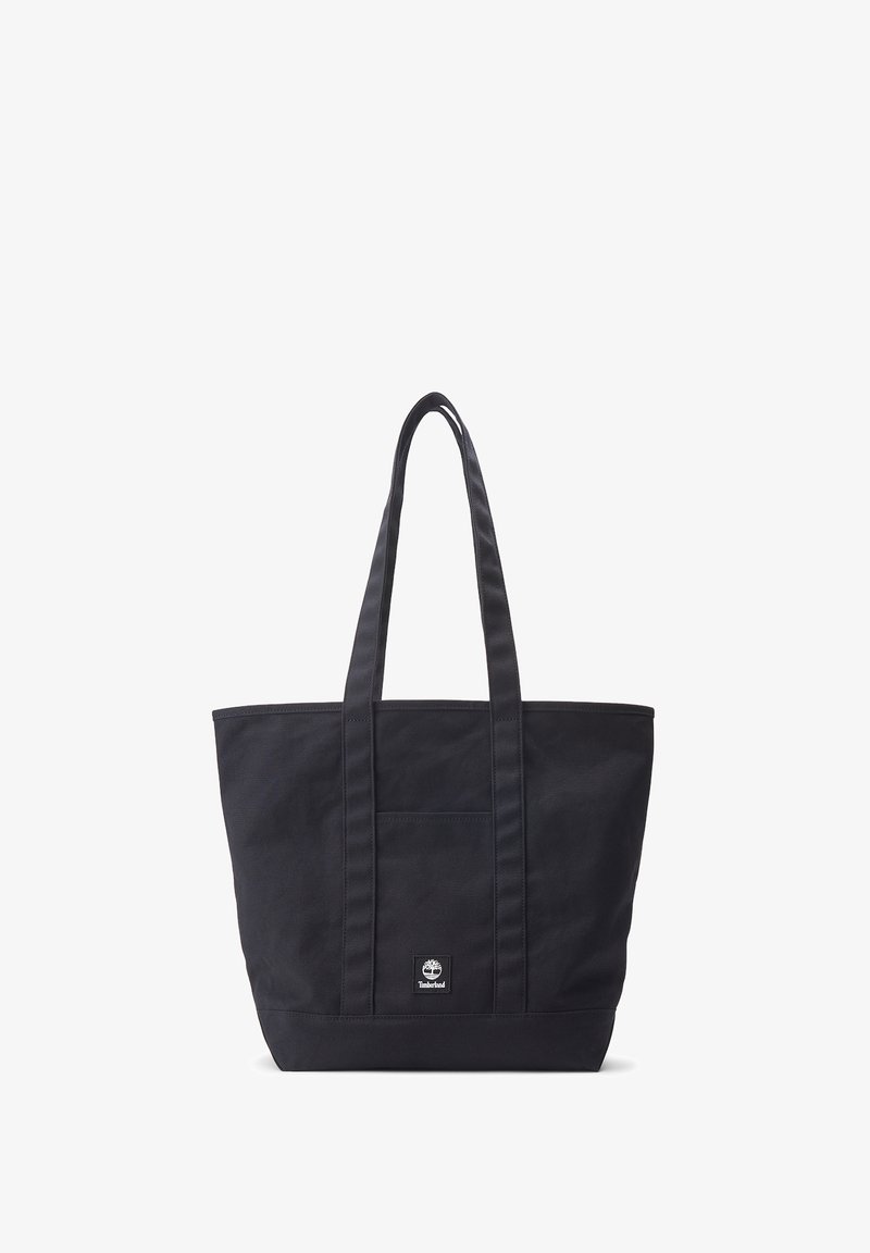 Svart canvas tote-väska med två långa handtag, en framficka och en liten logotypetikett nära basen. Enkel design, rymlig form.