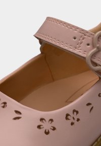 Chaussure synthétique rose avec un motif floral découpé, dotée d'une large bride maintenue par une boucle ronde. Texture lisse et design arrondi.