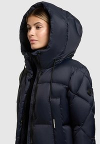 Marineblauwe pufferjas met een grote capuchon, voorzien van horizontale quiltlijnen, een zwarte rits en een logopatch op de linkermouw. Zachte, glanzende textuur.