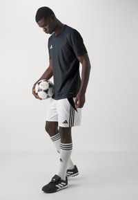 Idretts t-skjorte i svart, hvite shorts med tre svarte striper, svarte fotballsko, holder en svart og hvit fotball.