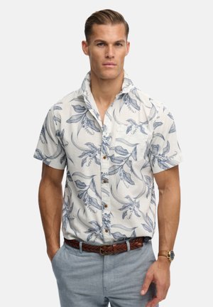 Mand med bagstrøget hår iført en hvid kortærmet skjorte med blå blomsterprint, lysegrå bukser, flettet brunt bælte og armbåndsur.