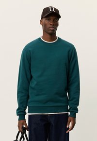 Πράσινη μπλούζα με στρογγυλή λαιμόκοψη, ριμπ μανσέτες και μικρό λογότυπο στο στήθος. Φορεμένη πάνω από λευκό πουκάμισο και συνδυασμένη με σκουρόχρωμο τζιν.