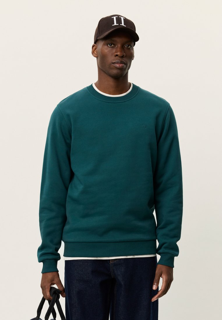 Πράσινη μπλούζα με στρογγυλή λαιμόκοψη, ριμπ μανσέτες και μικρό λογότυπο στο στήθος. Φορεμένη πάνω από λευκό πουκάμισο και συνδυασμένη με σκουρόχρωμο τζιν.