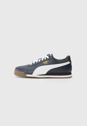 Marineblauwe Puma Roma sneaker met witte streep, witte veters, gumzool en gouden Puma Roma-logo aan de zijkant, weergegeven op een grijze achtergrond.