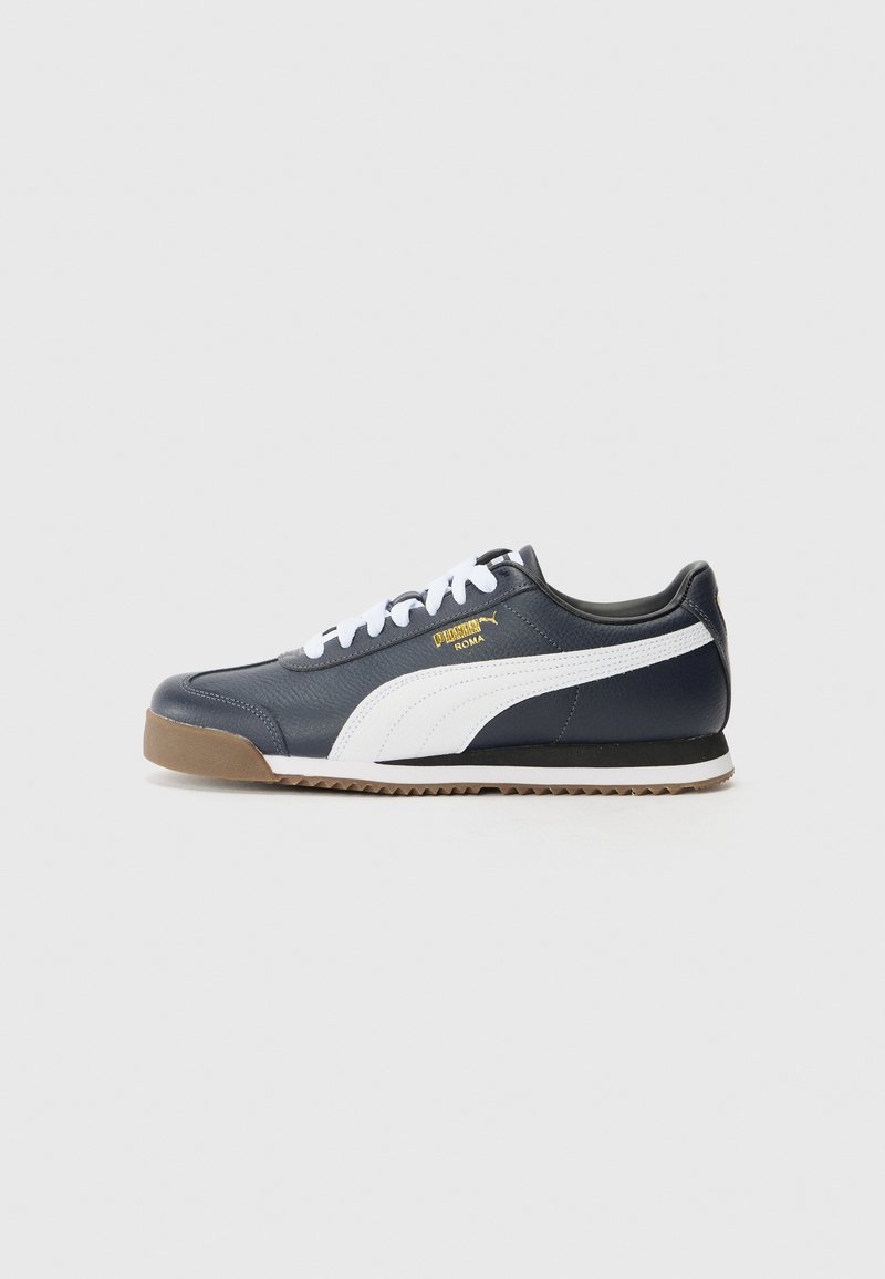 Baskets Puma Roma bleu marine avec bande blanche, lacets blancs, semelle en gomme, et logo Puma Roma doré sur le côté, présentées sur un fond gris.