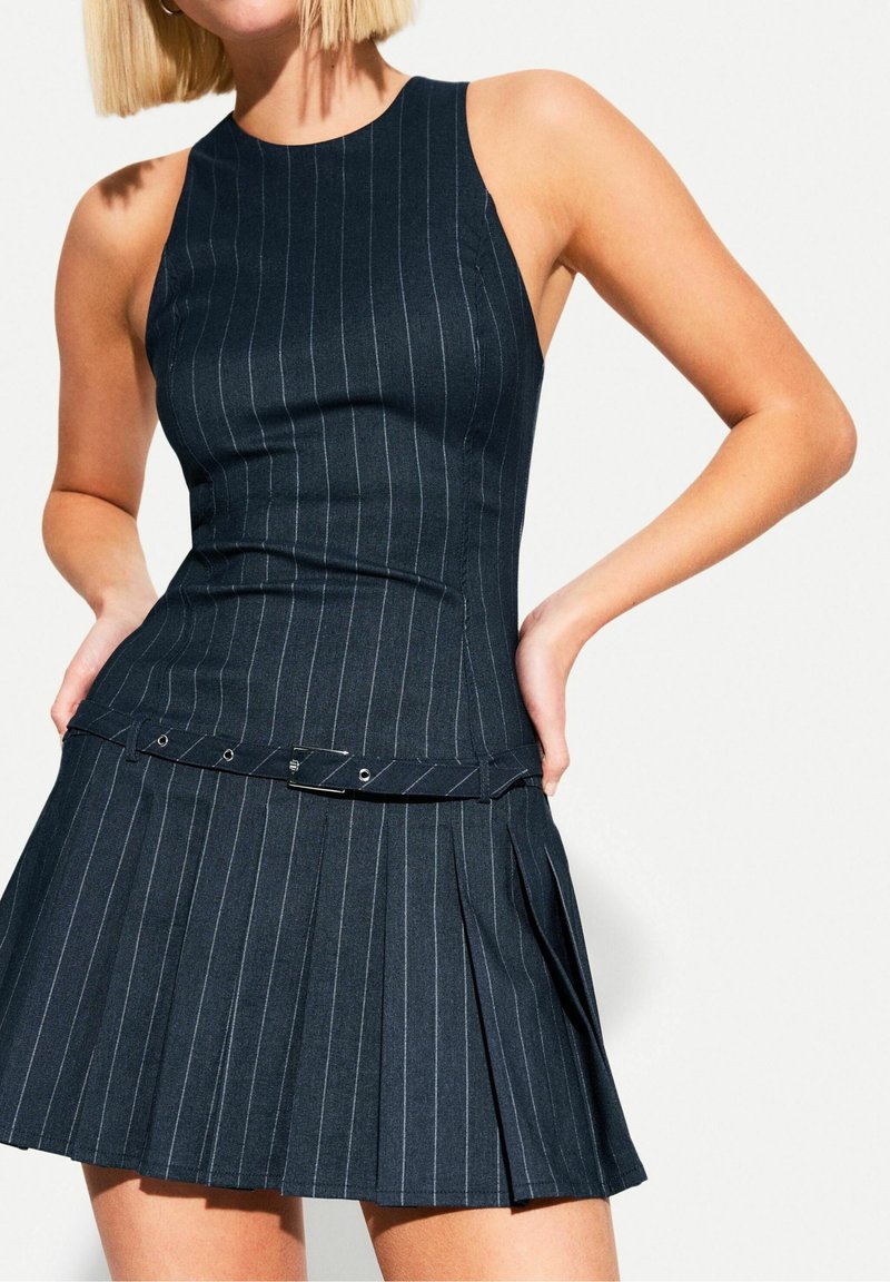 Femme portant une robe sans manches à fines rayures foncées avec un corsage ajusté, une jupe plissée et une ceinture assortie à la taille.