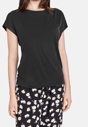 Femme portant un haut noir uni à manches courtes associé à un pantalon noir avec un motif floral marguerite blanche.