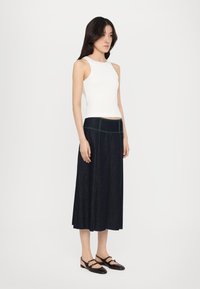 Hvitt ermeløst topp, mørk denim maxi skjørt med grønn søm, og brune sko med stroppe detaljer. Modellen står på en ensfarget bakgrunn.