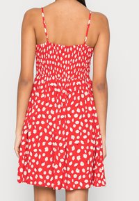 Červené šaty s bílými květinovými vzory, smockovaným živůtkem, tenkými ramínky a rozšířenou sukní. Textura podobná bavlně, ležérní letní design.