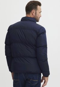 Marineblaue Steppjacke mit hohem Kragen, gestepptem Design und elastischen Bündchen. Sichtbarer Rückenteil und leichter Glanz in der Stoffstruktur.