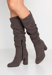 Bottes gris en daim au-dessus du genou avec un design affaissé, fermeture éclair sur le côté extérieur, et un talon bloc robuste. Texture lisse partout.