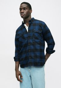 Camicia in flanella a quadri neri e blu navy con maniche lunghe, due tasche sul petto, bottoni sul davanti e polsini arrotolati. Indossata sopra una camicia bianca.