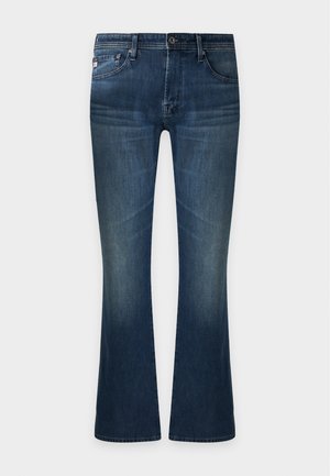 Homme portant un t-shirt blanc uni à manches courtes, un jean bleu et des chaussures noires, debout avec une main dans la poche.