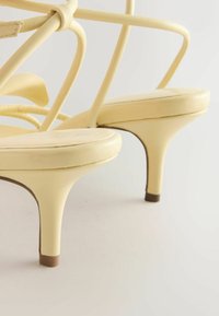 Sandales en cuir jaune à talons hauts avec fines brides à la cheville et talons aiguilles pointus sur une surface blanche unie.