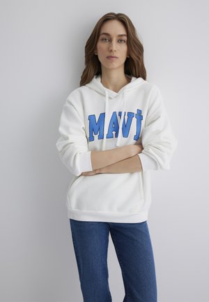 White hoodie with blue "MAVİ" text, drawstring, and front pocket. Paired with blue jeans, showcasing a relaxed fit and soft texture.
